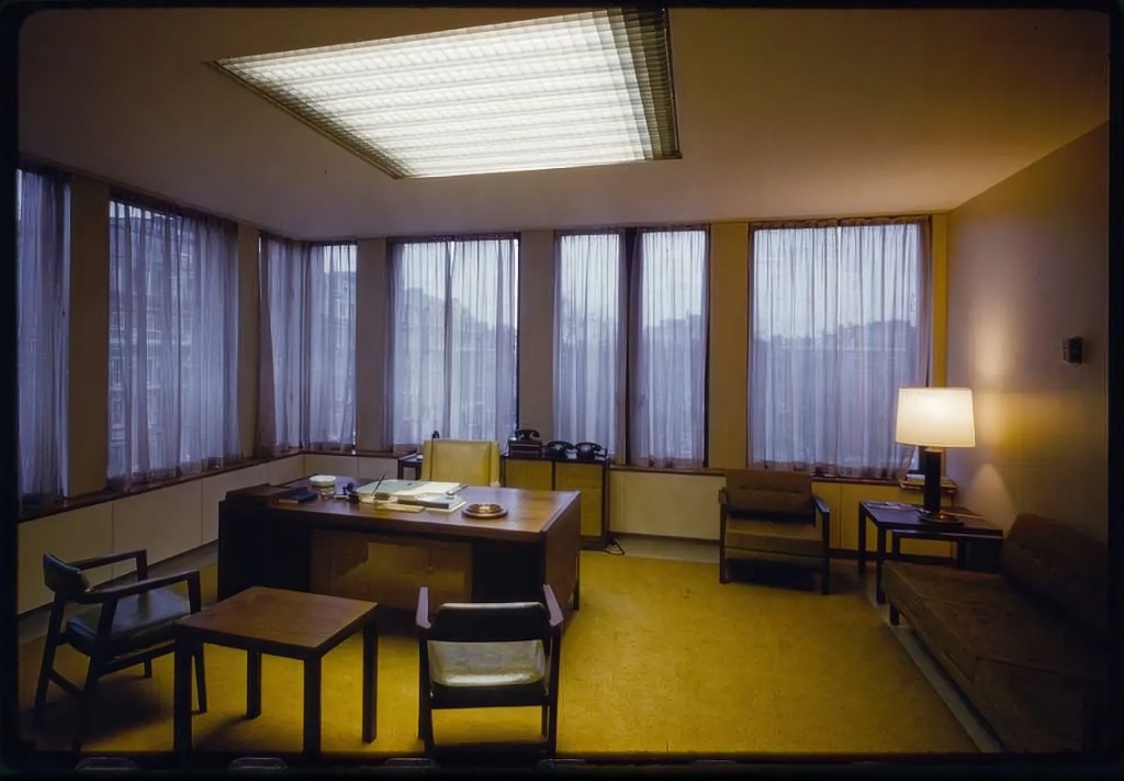 A 1960s office at dusk: a wooden desk with papers and an ashtray, chairs around a yellow carpet, and rain-grey city windows behind sheer curtains under a fluorescent ceiling light.